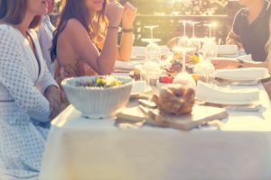 Group of young people eating outdoors. They are all happy, having fun, smiling laughing and talking. There are fruit and cheese platters on the table, some salad and a chicken or turkey. Back lit with lens flare.